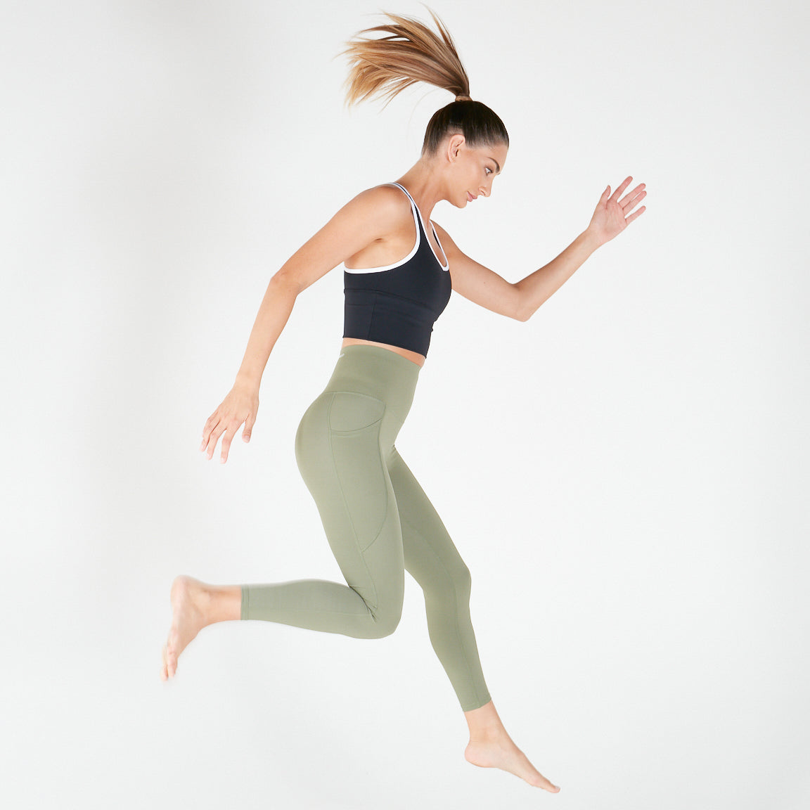 Woman in athletic wear mid-air wearing 7/8 Shaper Move Leggings in Olive on final sale