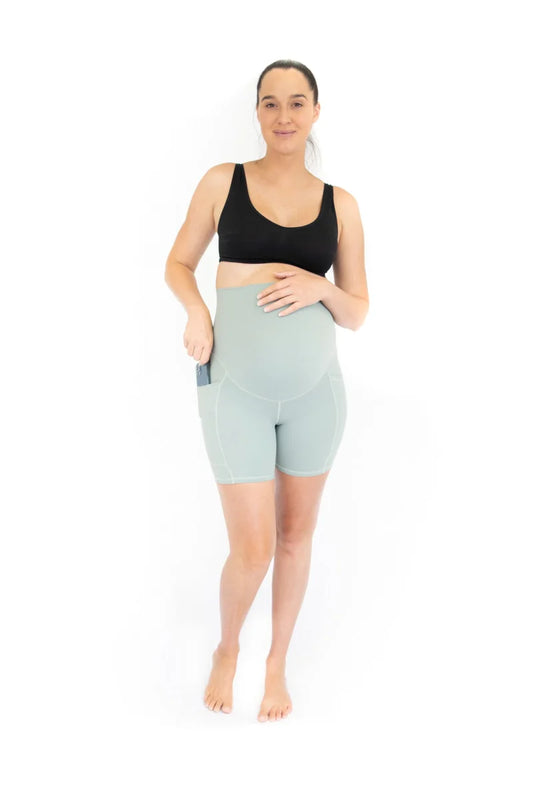 Woman in black sports bra and high-waisted mint green EmamaCo maternity bike shorts with pockets